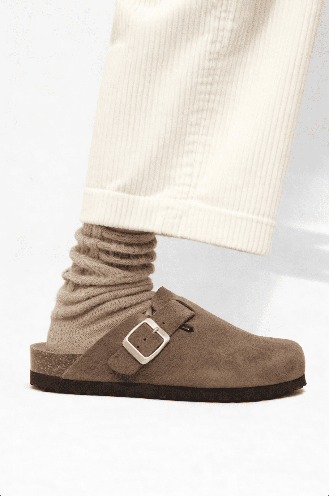 Zueco marrón con hebilla lateral llevado con calcetines beige, fotografiado de perfil sobre fondo neutro.