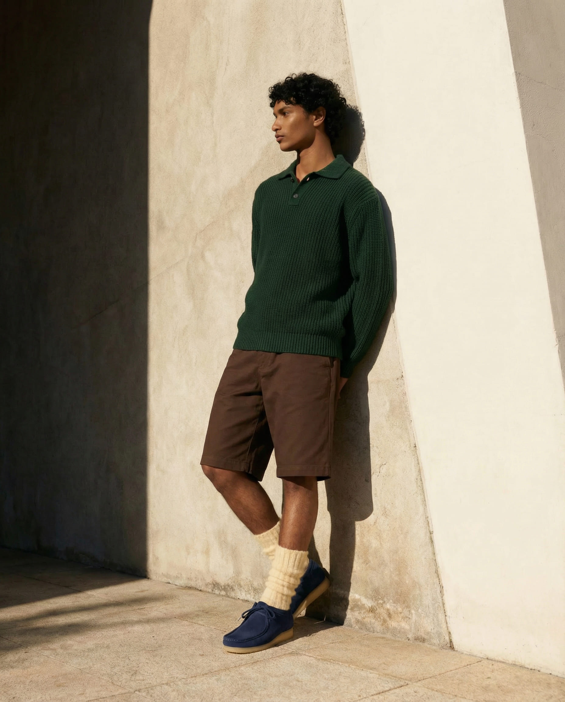 Persona de pie junto a una pared clara, con sudadera oscura y bermudas marrones, llevando zapatos oscuros de estilo casual.