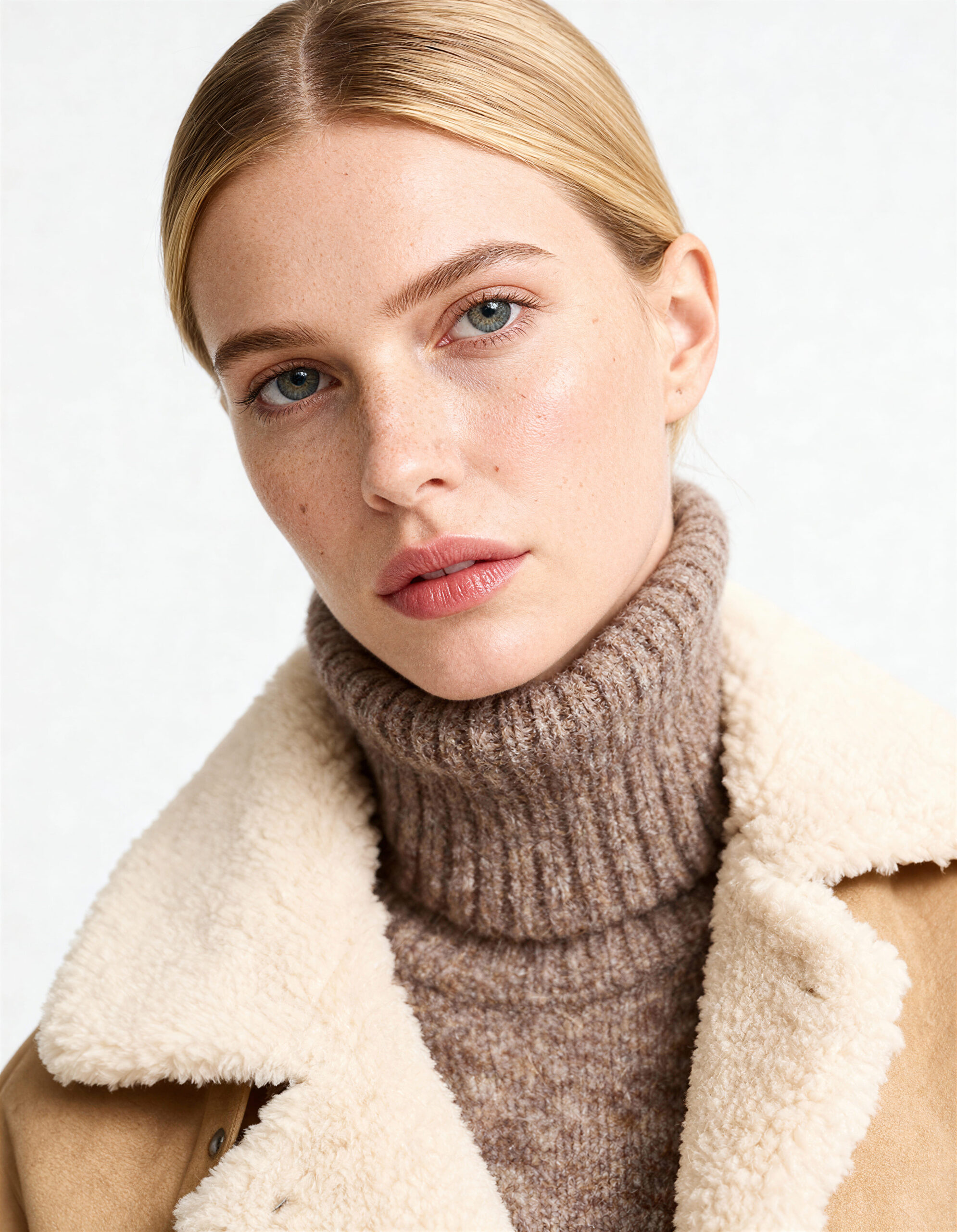 Retrato de una modelo con jersey de cuello alto y abrigo beige con interior de borreguito, sobre fondo neutro
