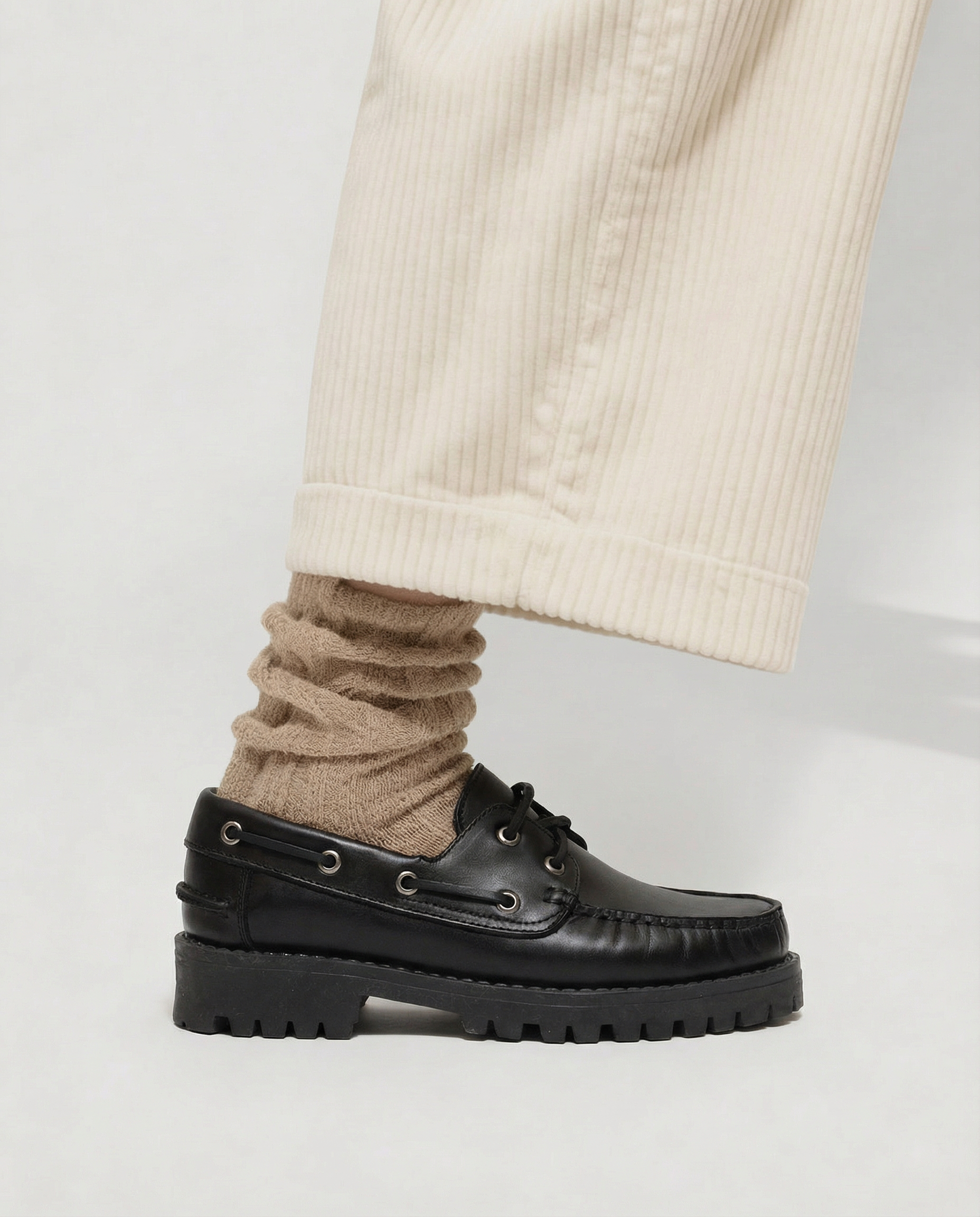 Zapato negro de suela gruesa llevado con calcetines beige y pantalón claro, fotografiado de perfil.